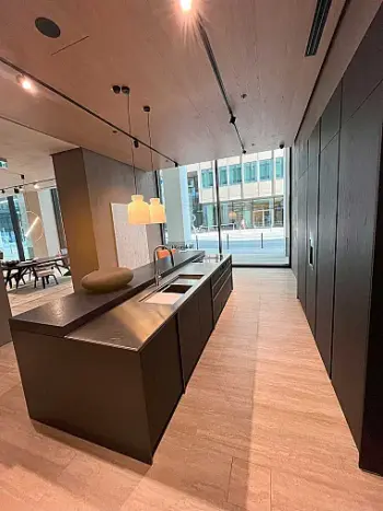 Molteni Abverkauf, Outlet, Angebot: Moderne Küche mit dunklen Schränken, Molteni&C-Insel mit eingebauter Spüle, Pendelleuchten und großen, raumhohen Fenstern mit Blick auf eine Stadtstraße. Das Design von Ratio Essenza Küche hebt hochwertige Küchengeräte hervor.