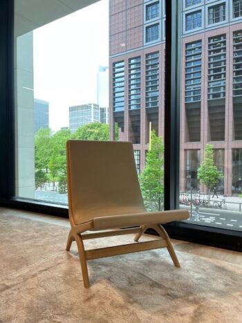 Ein moderner beiger Yoell Stuhl von Molteni&C steht auf einem Teppichboden neben großen Glasfenstern mit Blick auf hohe Gebäude und grüne Bäume.