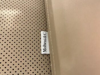 Nahaufnahme einer beigefarbenen perforierten Oberfläche mit einem Stoffetikett mit der Aufschrift Molteni&C, das teilweise unter einem glatten beigen Stoff versteckt ist und die raffinierten Details des Yoell Stuhl 2er Sets einfängt.