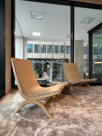 Zwei moderne beigefarbene Yoell Stuhl Loungesessel von Molteni&C sitzen als stilvolles 2er Set auf einem Plüschteppich in einem modernen Büroraum mit großen Fenstern und Blick auf benachbarte Gebäude.