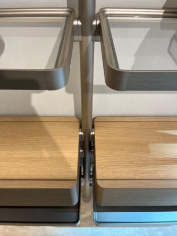 Zwei Regale aus dem Hector Regalsystem von Molteni&C sind an einer Wand montiert; die oberen Regale sind aus Glas, die unteren aus hellem Holz, alle getragen von einer zentralen Metallstange.