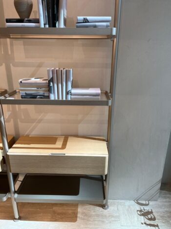 Ein Regal von Molteni&C Hector Regalsystem beherbergt mehrere übersichtlich angeordnete Bücher, eine geschlossene Aufbewahrungsbox und eine Vase. Das Regal wirft einen Schatten auf die helle Wand und den Boden.