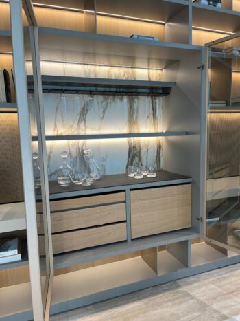 Eine moderne Molteni&C-Glasvitrine mit offenen Türen stellt Glaskaraffen, Gläser und Regale aus. Sie ist mit einer Rückwand in Marmoroptik und Holzschubladen aus dem 505 UP System ausgestattet.