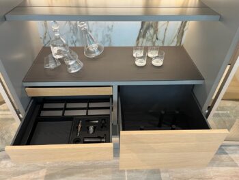 Eine Molteni&C 505 UP System-Arbeitsplatte mit Glaskaraffen und Gläsern über zwei offenen Schubladen - eine mit unterteilten Fächern und Barutensilien, die andere leer - vor einer markanten Marmorplatte.