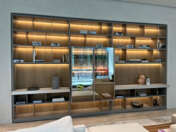 Moderne Wandeinbauregale aus dem Molteni&C 505 UP System mit integrierter Beleuchtung präsentieren Bücher, Dekorationsgegenstände und einige Spiegel. Eine Straßenansicht mit einem roten Auto ist durch eine Fensterspiegelung sichtbar.