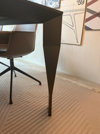 Eine Nahaufnahme eines modernen schwarzen Diamond Designtischs und eines braunen Bürostuhls auf einem strukturierten beigen Teppich, mit gerahmten Kunstwerken an der Wand im Hintergrund, die die raffinierte Ästhetik von Molteni&C widerspiegeln.