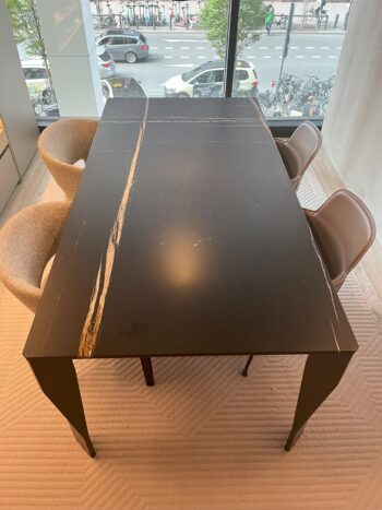 Rechteckiger schwarzer Designtisch mit einem hellen Streifen, umgeben von zwei gepolsterten Stühlen und zwei braunen Stühlen, in der Nähe eines großen Fensters mit Blick auf eine Straße mit Autos und Fahrrädern - ein raffinierter Molteni&C-Touch.