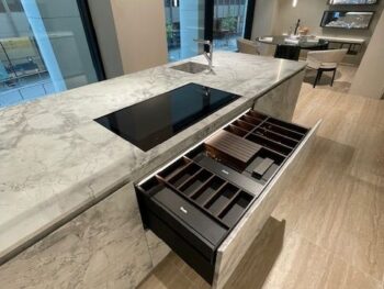 Molteni Abverkauf, Outlet, Angebot: Offene Küchenschublade mit geordneten Fächern unter einer Marmorarbeitsplatte von Molteni&C, neben einem schwarzen Elektrokochfeld, in der modernen Küche Intersection mit hellem Holzfußboden.