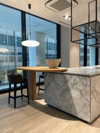Molteni Abverkauf, Outlet, Angebot: Moderne Küche mit einer Marmorinsel von Molteni&C Intersection, einem Holztisch, zwei schwarzen Stühlen, einer Hängelampe und einer dekorativen Schale - alles vor großen Fenstern mit Blick auf Bürogebäude.