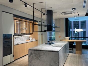 Abverkauf, Outlet, Angebot: Moderne Küche mit einer großen Marmorinsel aus der Kollektion Intersection von Molteni&C, minimalistischen Schränken aus hellem Holz, Einbaugeräten und geometrischen schwarzen Deckenleuchten. Großes Fenster und Esstisch im Hintergrund.