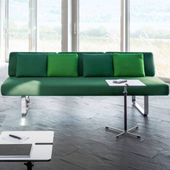 Ein grünes Joline-Sofa mit vier passenden Kissen steht vor großen Fenstern; ein kleiner weißer Beistelltisch mit einer Fernbedienung steht daneben, und auf einem zweiten Tisch im Vordergrund liegen ein Notizblock und ein Stift.