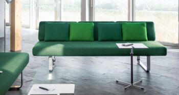 Ein modernes grünes Girsberger Joline-Sofa mit fünf grünen Kissen steht vor großen Fenstern; ein kleiner Beistelltisch und ein Notizblock stehen daneben auf einem grauen Fliesenboden.
