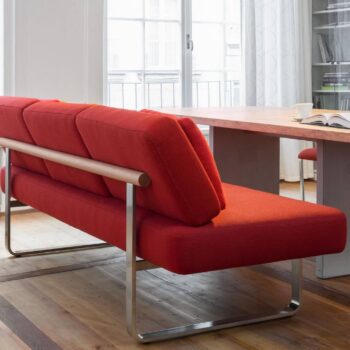 Ein modernes rotes Joline-Sofa von Girsberger mit einem schlanken Metallrahmen steht neben einem Holztisch in einem hellen Raum mit Holzböden, großen Fenstern und Bücherregalen.