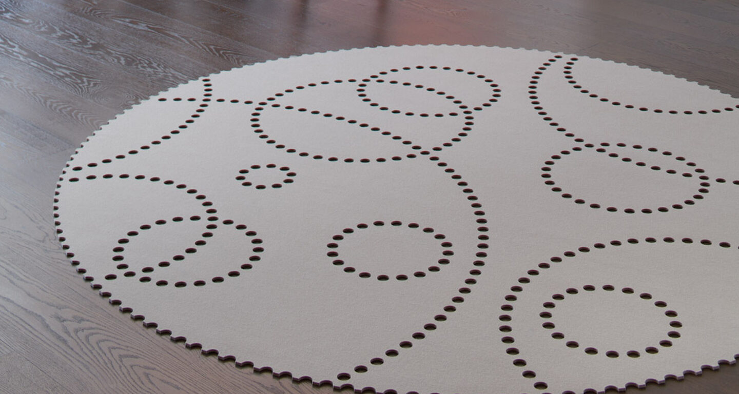 Ein runder, heller Hey-Sign-Teppich mit einem Muster aus verschieden großen Kreisen aus kleinen Perforationen, der auf einem dunklen Holzboden liegt.