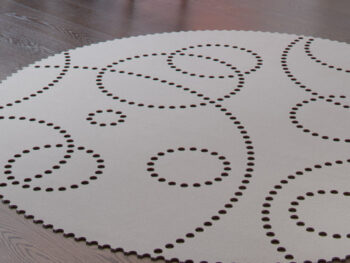 Ein runder, heller Hey-Sign-Teppich mit einem Muster aus verschieden großen Kreisen aus kleinen Perforationen, der auf einem dunklen Holzboden liegt.