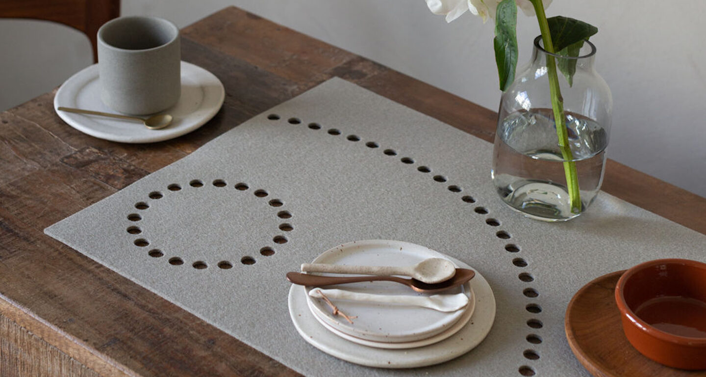 Ein ordentlich gedeckter Tisch mit Keramiktellern, -tassen und -löffeln auf einer hölzernen Unterlage, daneben eine Vase mit einer weißen Blume und ein Glas Wasser mit einem Hey-Sign-Platzset mit kreisförmigen Ausschnitten.