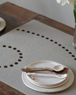 Ein ordentlich gedeckter Tisch mit Keramiktellern, -tassen und -löffeln auf einer hölzernen Unterlage, daneben eine Vase mit einer weißen Blume und ein Glas Wasser mit einem Hey-Sign-Platzset mit kreisförmigen Ausschnitten.