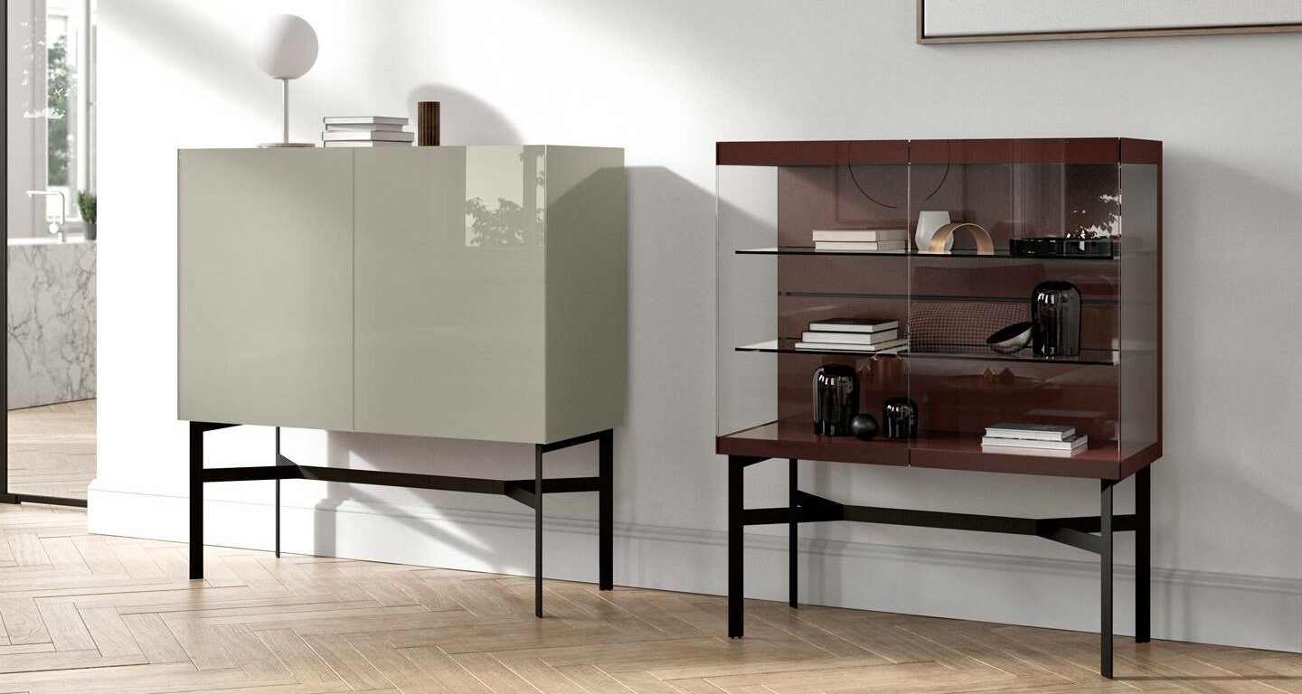 Zwei moderne Schränke auf schwarzen Metallbeinen stehen nebeneinander; der eine ist geschlossen und in glänzendem Beige gehalten, der andere offen mit Glastüren, die dekorative Gegenstände zeigen.