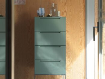 Eine hohe, mintgrüne Mell-Kommode mit einer Messinglampe, einer Glasflasche und kleinen Gegenständen darauf steht an einer Holzwand neben einer Glastür.