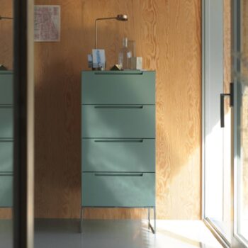 Eine hohe, mintgrüne Mell-Kommode mit einer Messinglampe, einer Glasflasche und kleinen Gegenständen darauf steht an einer Holzwand neben einer Glastür.