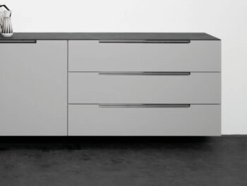 Modernes Interlübke-Sideboard mit drei horizontalen Schubladen und einer glatten schwarzen Arbeitsplatte vor einer weißen Wand, auf der ein Teil eines gerahmten Kunstwerks zu sehen ist.