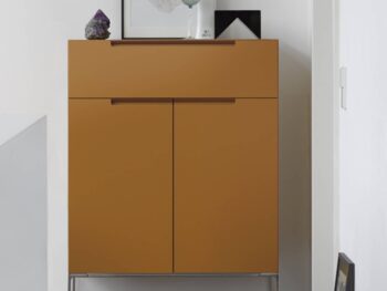 Senfgelbes Interlübke Sideboard mit zwei Türen und zwei Schubladen, auf Metallfüßen stehend, bestückt mit Dekorationsobjekten und Kunstwerken, vor einer weißen Wand.