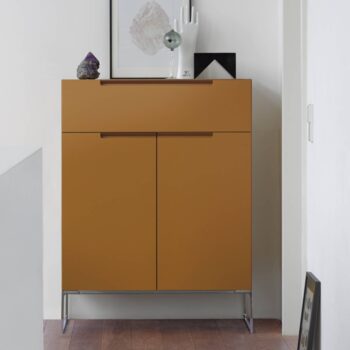 Senfgelbes Interlübke Sideboard mit zwei Türen und zwei Schubladen, auf Metallfüßen stehend, bestückt mit Dekorationsobjekten und Kunstwerken, vor einer weißen Wand.