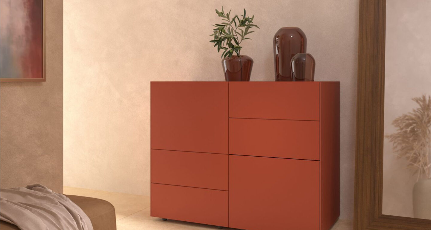 Ein roter Schrank mit klaren Linien steht vor einer beigen Wand, darauf eine Topfpflanze und zwei braune Ziervasen, daneben ein großer Spiegel.