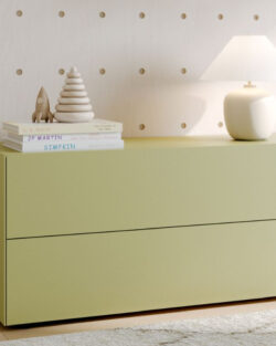 Ein hellgrüner Schrank mit zwei Schubladen steht an einer Wand aus Steckbrettern, darauf eine Lampe, gestapelte Bücher und kleine dekorative Gegenstände.