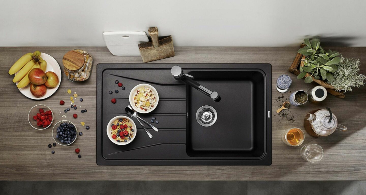 Draufsicht auf eine Küchentheke mit einer schwarzen Spüle, zwei Schalen mit Müsli und Obst, frischem Obst, einer Teekanne, Tassen und Schneidebrettern, die um die Spüle herum angeordnet sind.