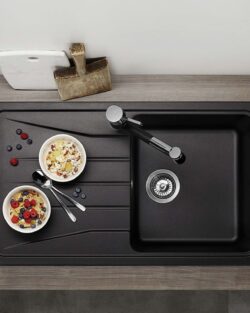 Draufsicht auf eine Küchentheke mit einer schwarzen Spüle, zwei Schalen mit Müsli und Obst, frischem Obst, einer Teekanne, Tassen und Schneidebrettern, die um die Spüle herum angeordnet sind.