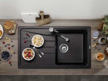 Draufsicht auf eine Küchentheke mit einer schwarzen Spüle, zwei Schalen mit Müsli und Obst, frischem Obst, einer Teekanne, Tassen und Schneidebrettern, die um die Spüle herum angeordnet sind.