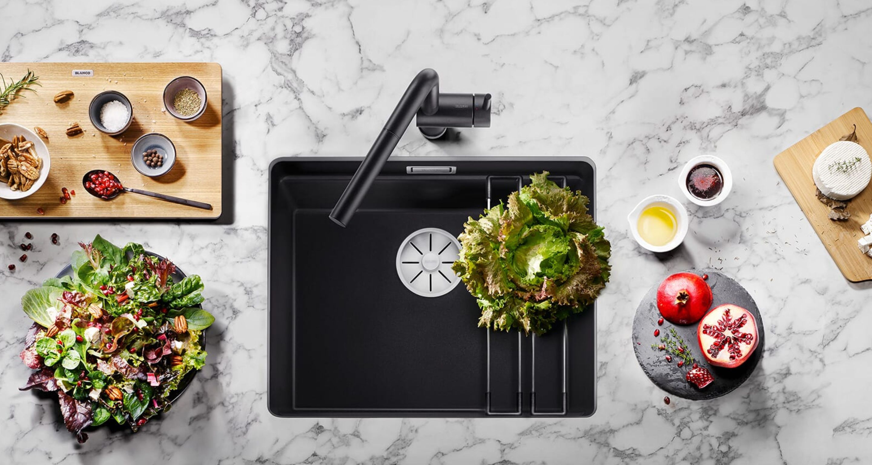 Draufsicht auf eine moderne schwarze Küchenspüle mit einem Salat, frischem Gemüse, Granatapfel und Gewürzzutaten auf einer Marmorarbeitsplatte.