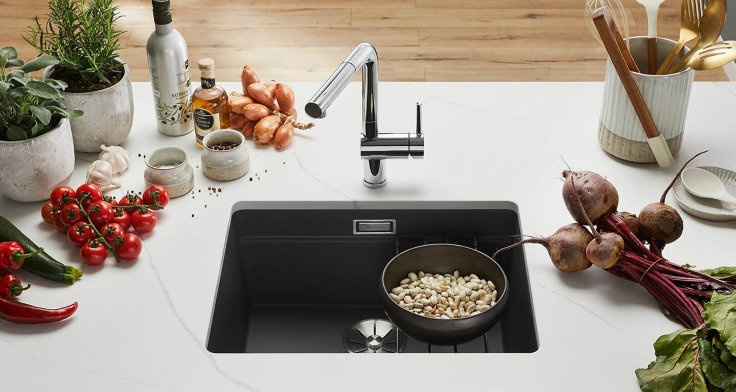 Eine moderne Küchenspüle mit einer Schale weißer Bohnen im Becken, umgeben von frischem Gemüse, Kräutern und Küchenutensilien auf einer weißen Arbeitsplatte.