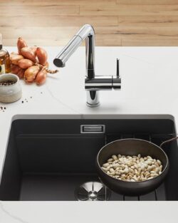 Eine moderne Küchenspüle mit einer Schale weißer Bohnen im Becken, umgeben von frischem Gemüse, Kräutern und Küchenutensilien auf einer weißen Arbeitsplatte.