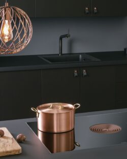 Moderne Küche mit schwarzen Schränken, einem Bora-Induktionsherd, auf dem ein Kupfertopf steht, einem hölzernen Schneidebrett mit Kastanien und einer geometrischen Pendelleuchte über dem Kopf.