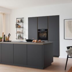 Moderne Küche mit dunklen Schränken, Inselarbeitsplatte, eingebautem Ofen, großen Fenstern, Topfpflanze und Essbereich mit minimalistischem Dekor.