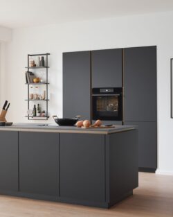Moderne Küche mit dunklen Schränken, Inselarbeitsplatte, eingebautem Ofen, großen Fenstern, Topfpflanze und Essbereich mit minimalistischem Dekor.