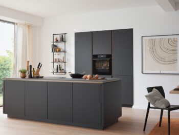 Moderne Küche mit dunklen Schränken, Inselarbeitsplatte, eingebautem Ofen, großen Fenstern, Topfpflanze und Essbereich mit minimalistischem Dekor.
