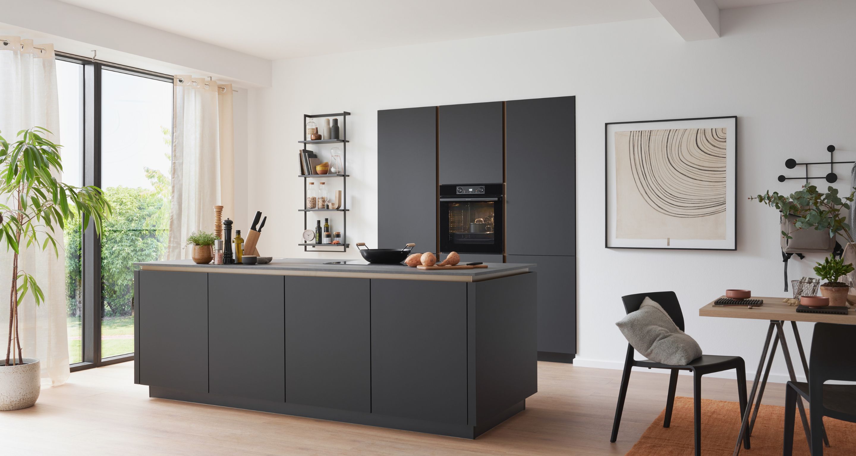 Moderne Küche mit dunklen Schränken, Inselarbeitsplatte, eingebautem Ofen, großen Fenstern, Topfpflanze und Essbereich mit minimalistischem Dekor. Moderne Küche mit dunklen Schränken, Inselarbeitsplatte, eingebautem Ofen, großen Fenstern, Topfpflanze und Essbereich mit minimalistischem Dekor.