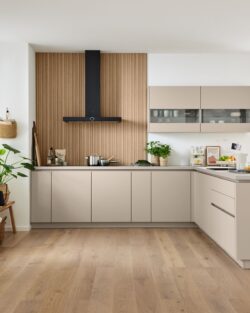 Moderne Küche mit beigen Schränken, Holzakzenten, eingebautem Ofen, Pflanzen und einem Esstisch in der Nähe großer Fenster, die viel Tageslicht hereinlassen.