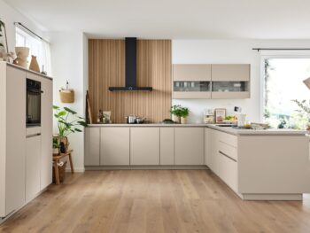 Moderne Küche mit beigen Schränken, Holzakzenten, eingebautem Ofen, Pflanzen und einem Esstisch in der Nähe großer Fenster, die viel Tageslicht hereinlassen.