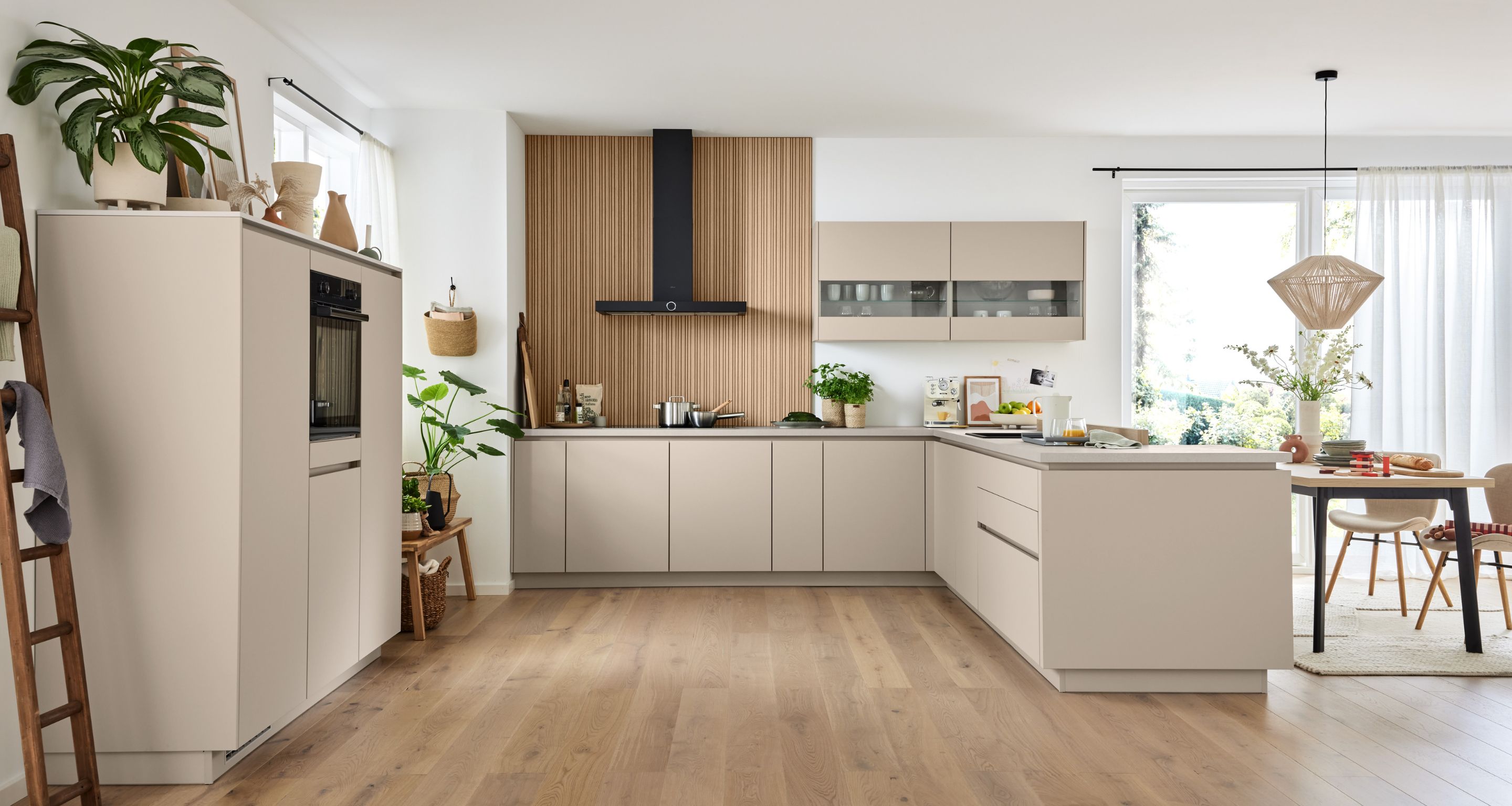 Moderne Küche mit beigen Schränken, Holzakzenten, eingebautem Ofen, Pflanzen und einem Esstisch in der Nähe großer Fenster, die viel Tageslicht hereinlassen. Moderne Küche mit beigen Schränken, Holzakzenten, eingebautem Ofen, Pflanzen und einem Esstisch in der Nähe großer Fenster, die viel Tageslicht hereinlassen.