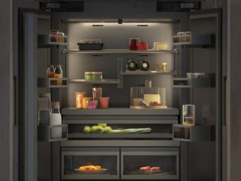 Ein offener Kühlschrank mit ordentlich angeordneten Flaschen, Gläsern, Obst, Gemüse, Eiern und Getränken auf Regalen und in Schubladen.