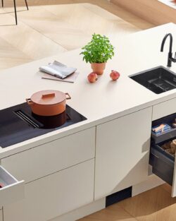 Moderne Küche mit einer weißen Insel mit Induktionskochfeld, Spüle, Kupfertopf, Kräutertopf und zwei offenen Schubladen für Küchenutensilien.