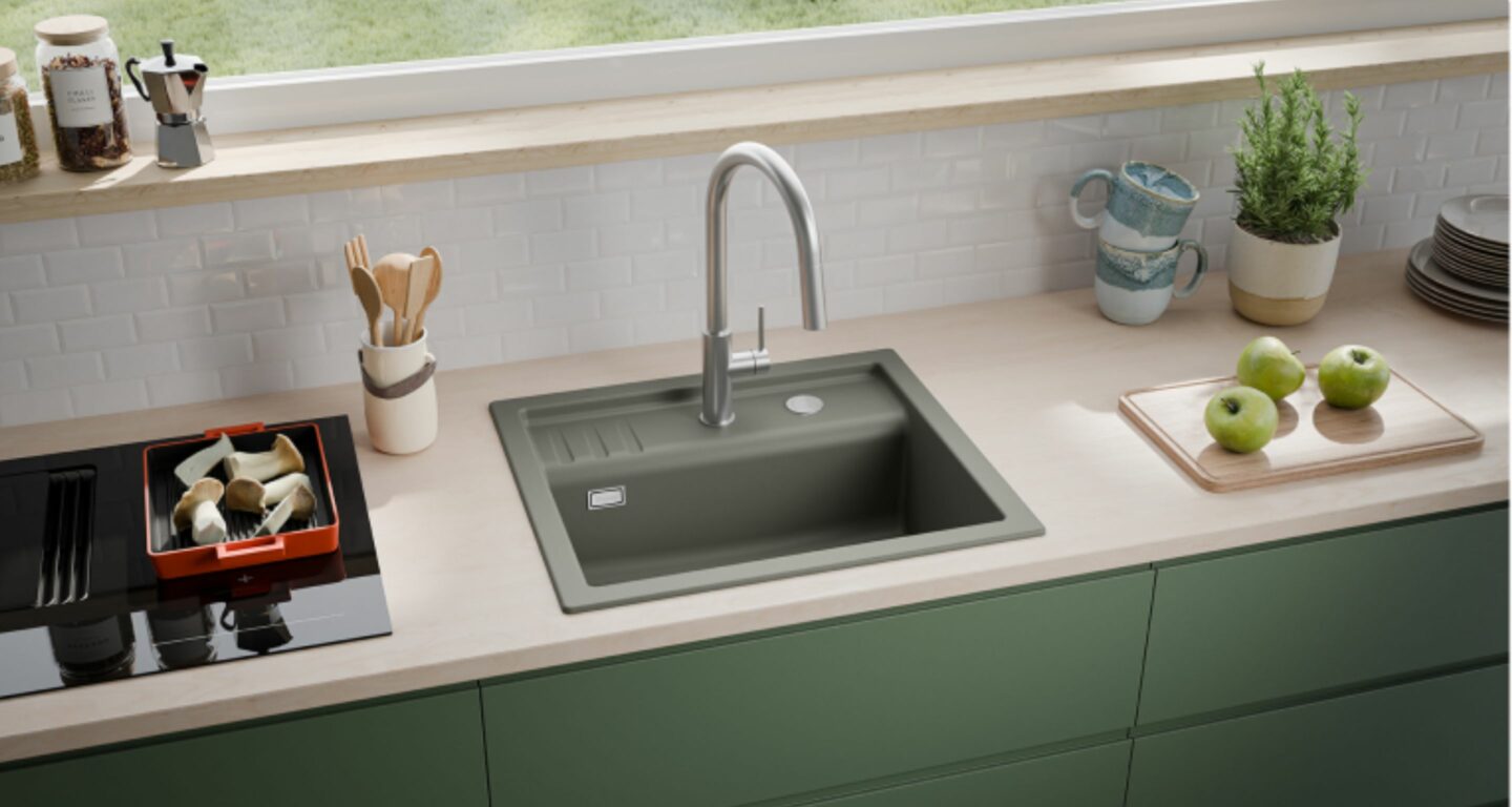 Eine moderne Küchenarbeitsplatte mit einer grauen, quadratischen Spüle, einem hohen Wasserhahn, grünen Schränken, Utensilien, Äpfeln, Tassen und Pflanzen, vor einem Fenster mit Blick auf das Gras draußen.