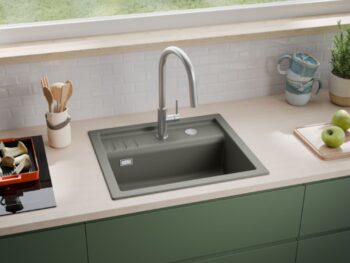 Eine moderne Küchenarbeitsplatte mit einer grauen, quadratischen Spüle, einem hohen Wasserhahn, grünen Schränken, Utensilien, Äpfeln, Tassen und Pflanzen, vor einem Fenster mit Blick auf das Gras draußen.
