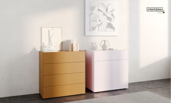 Zwei moderne Kommoden vom Möbelhaus Bad Homburg, eine senfgelbe mit vier Schubladen und eine hellrosa mit zwei Schubladen und einem Schrank, stehen nebeneinander vor einer weißen Wand mit minimalem Dekor darauf.
