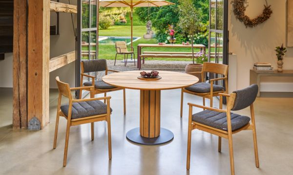 In einem Raum, dessen offene Türen zu einem Garten und einem Poolbereich im Freien führen, steht ein runder Holztisch mit vier gepolsterten Stühlen, die aus dem Möbelhaus Bad Homburg stammen. In einem Raum, dessen offene Türen zu einem Garten und einem Poolbereich im Freien führen, steht ein runder Holztisch mit vier gepolsterten Stühlen, die aus dem Möbelhaus Bad Homburg stammen.