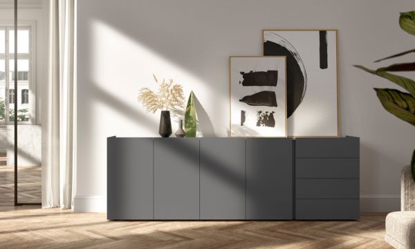 Ein modernes Sideboard vom Möbelhaus Bad Homburg mit grauen Schränken ist mit abstrakter gerahmter Kunst, Keramikvasen und getrockneten Pflanzen dekoriert und steht vor einer weißen Wand in einem sonnendurchfluteten Raum.